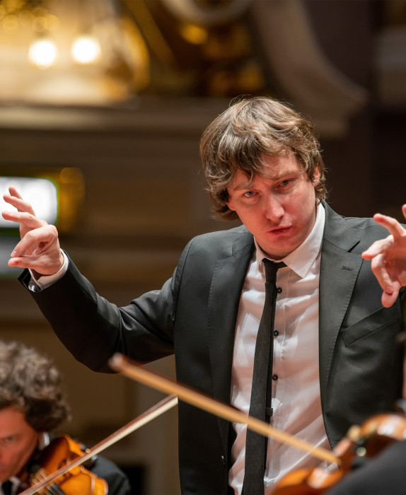 Maxim conducts the orchestra. String players are in shot.