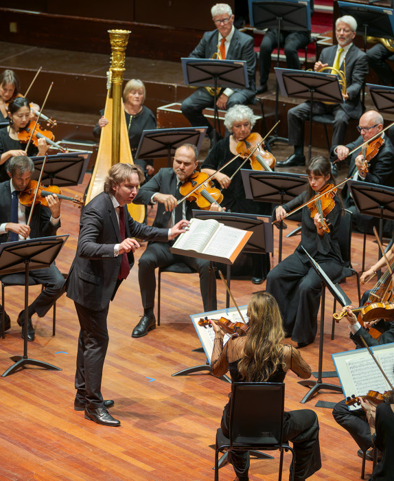 Maxim conducts the orchestra from the Usher Hall. He wears a black suit.