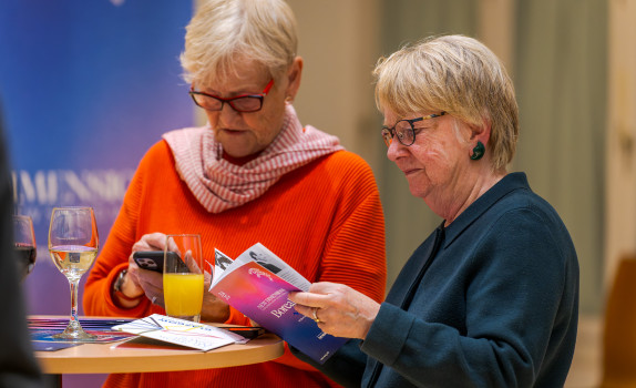 Two audience members read a programme. They are older women, one wears a red jumper and scarf, the other has a blue jumper on. Both wear glasses.