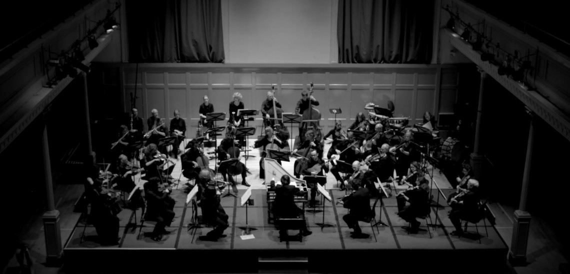 A shot from the gallery in the Queen's Hall of the orchestra in black and white. Maxim Emelyanychev is conducting while sitting at the harpsichord.