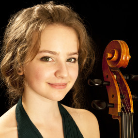 A woman in a dark dress looks towards the camera at a slight angle. She looks with a closed smile and is against a back background. The scroll of a cello is visible.