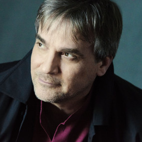 A man in a dark coat looks off to the right. Light shines on his face casting a slight shadow on his left hand side. He is against a dark background