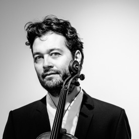 A man looks to the camera with a slightly cocked head. A viola scroll is visible and obscures the side of the man's face