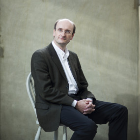 Man in a black suit and white shirt sits on on a white share against a cement wall background. The man looks at the camera and clasps his hands.