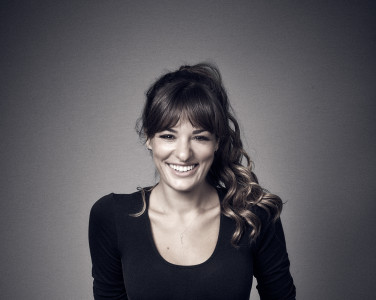 Woman stands against a lit background. her hand are in her pockets and she smiles directly at the camera