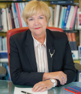Professor Pamela Gillies sitting at a desk with her hands resting on it, smiling at the camera.