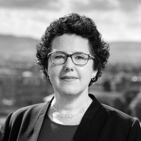 Black and white headshot of Joanna Baker. She looks directly at the camera, smiling, wearing glasses and a blazer.