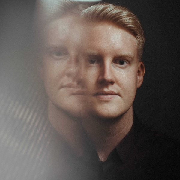 A man looks towards the camera against a black background. The left side of the image has been distorted and a faint copy of the man's face is visible.