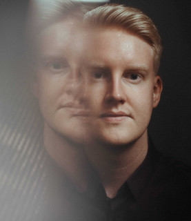 A man looks towards the camera against a black background. The left side of the image has been distorted and a faint copy of the man's face is visible.