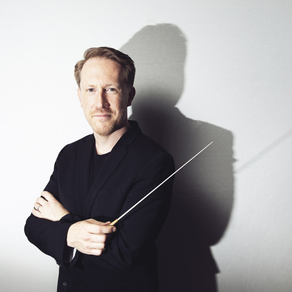 Conductor Simon Crawford-Phillips poses with arms crossed, holding the baton, looking directly into the camera.