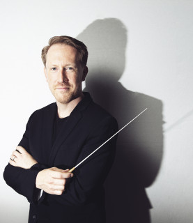Conductor Simon Crawford-Phillips poses with arms crossed, holding the baton, looking directly into the camera.
