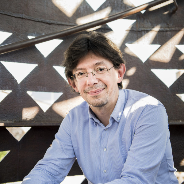Conductor Václav Luks sits leaning forward, arms resting on his knees, smiling as he looks at the camera.