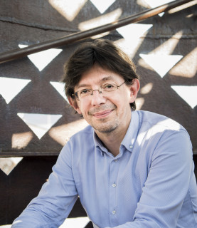 Conductor Václav Luks sits leaning forward, arms resting on his knees, smiling as he looks at the camera.