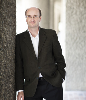 Conductor Andrew Manze stands in a suit, facing the camera, leaning against a wall to his right.