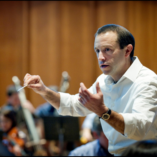 Conductor Mark Wigglesworth, captured from the side mid-conducting, his arms raised with a baton held in his right hand.