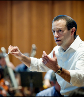 Conductor Mark Wigglesworth, captured from the side mid-conducting, his arms raised with a baton held in his right hand.
