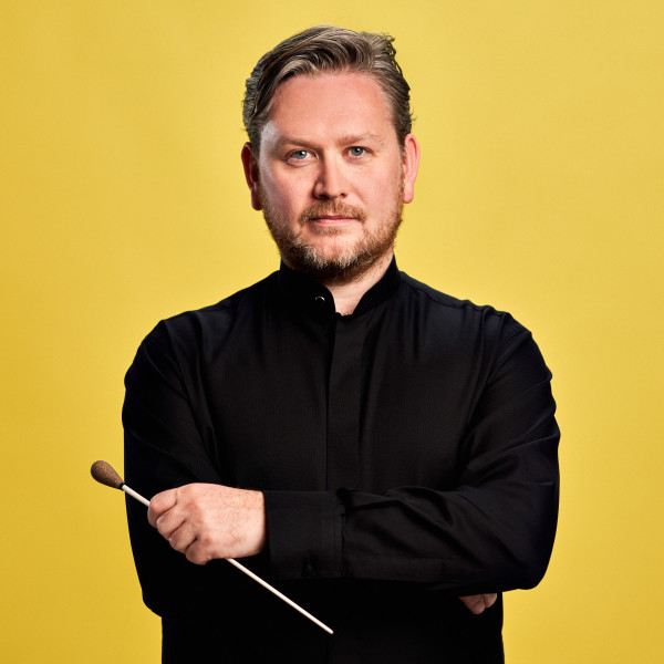 Conductor Jonathan Bloxham stands with his arms crossed, looking into the camera with a light smile, in front of a yellow background.