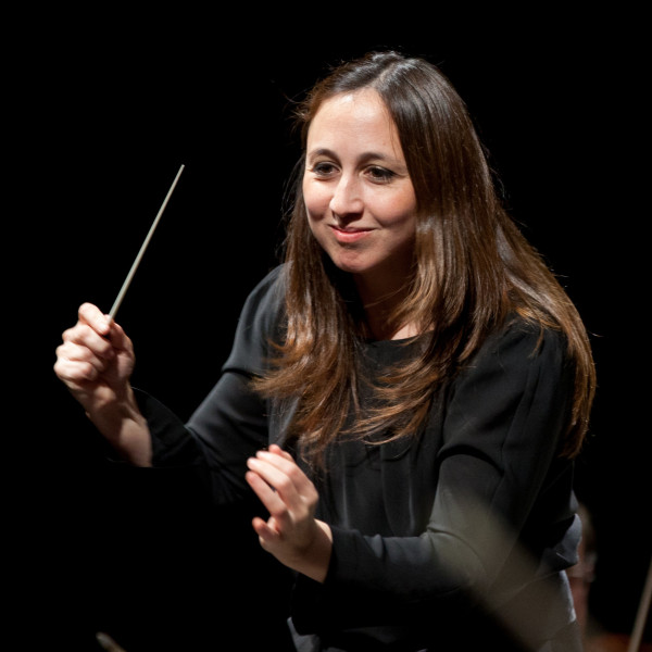 Conductor Joana Carneiro smiles as she leads the orchestra, her hand raised mid-gesture.
