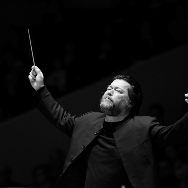 Conductor Joseph Swensen is captured in black and white, mid-conducting with both arms raised in the air, eyes closed in concentration.