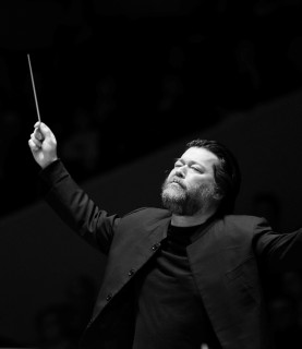 Conductor Joseph Swensen is captured in black and white, mid-conducting with both arms raised in the air, eyes closed in concentration.