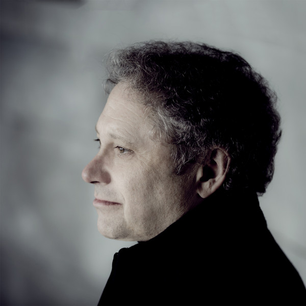 A man in a dark outdoor jacket looks off to the left with light shining on his face. He is against a dark stone background