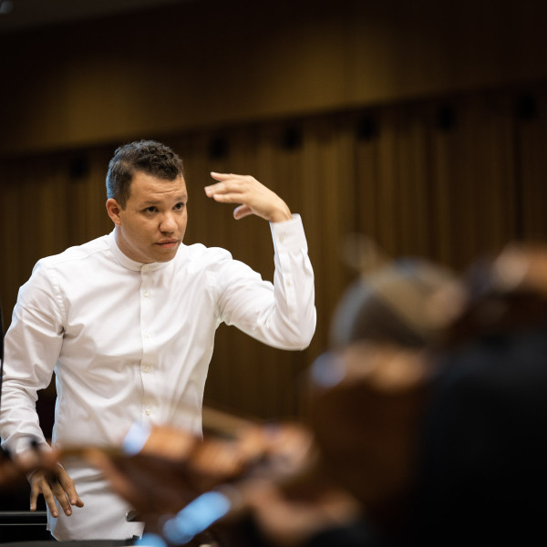 Conductor Ryan Bancroft directs the orchestra, his left arm raised, in a dynamic conducting pose.