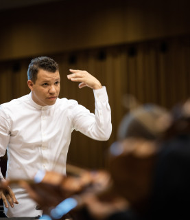 Conductor Ryan Bancroft directs the orchestra, his left arm raised, in a dynamic conducting pose.