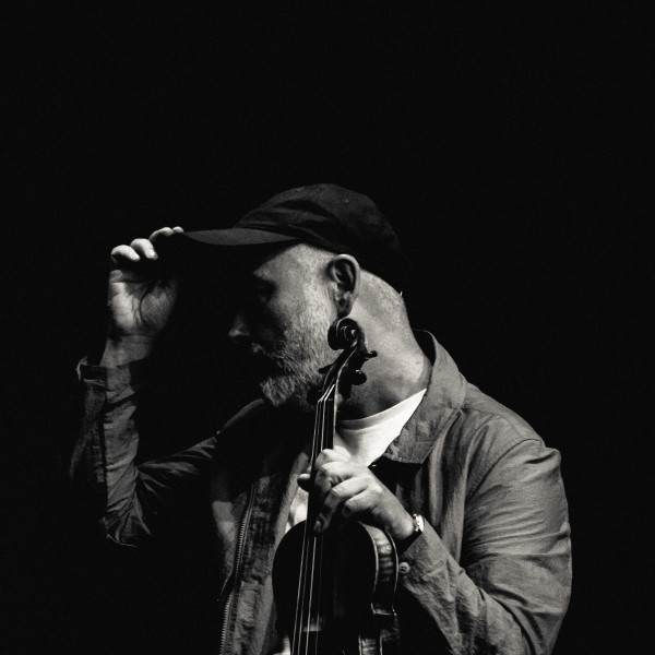 Aidan O'Rourke sits in a dark room with a fiddle. The picture is in black and white.