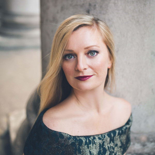 Soprano Lucy Crowe poses sitting down in an elegant dress, looking directly at the camera.