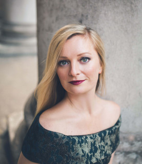 Soprano Lucy Crowe poses sitting down in an elegant dress, looking directly at the camera.