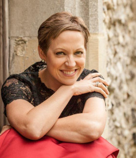 Soprano Julia Doyle sits, smiling with crossed arms, her right hand touching her left shoulder, chin resting on her hand.
