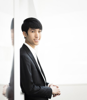 Pianist Eric Lu stands, leaning against a white wall, facing the camera to his right.