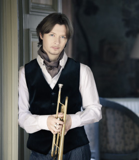 Trumpeter Sergei Nakariakov leans in a doorway, holding his trumpet in front of him.
