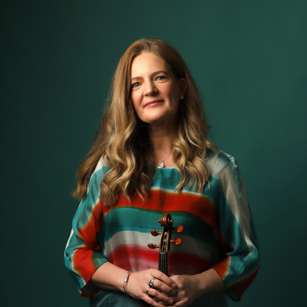 Violinist Rachel Podger stands, smiling at the camera, holding her violin in front of her.