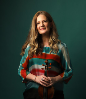 Violinist Rachel Podger stands, smiling at the camera, holding her violin in front of her.