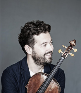 Violinist Lawrence Power sits, smiling and looking to his left, holding his violin close to his chest.