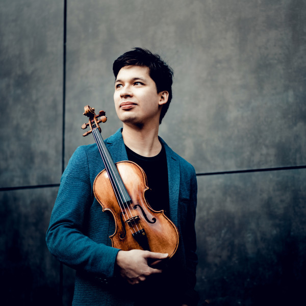 Violinist Aylen Pritchin stands, looking into the distance towards his right, holding his violin in front of him.