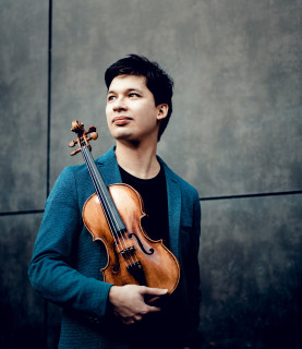 Violinist Aylen Pritchin stands, looking into the distance towards his right, holding his violin in front of him.