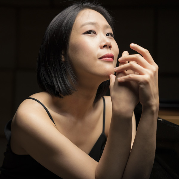 Pianist Yeol Eum Son poses leaning against a piano, her elbows resting on its edge, one hand cradling the other in mid-air, while she tilts her head back and gazes upwards.