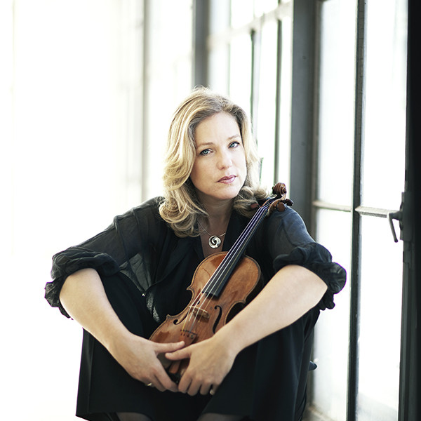 Violinist Isabelle van Keulen sits on the floor with legs angled, arms resting on her knees, holding her violin between her legs.