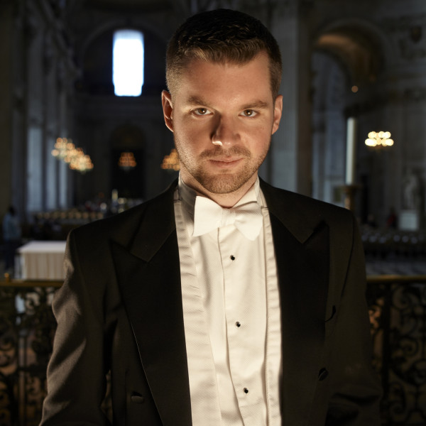 Bass-baritone Edward Grint stands, head slightly tilted down, gazing directly up at the camera.