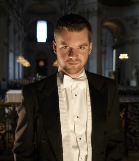 Bass-baritone Edward Grint stands, head slightly tilted down, gazing directly up at the camera.