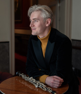Oboist Ivan Podyomov sits at a small table, his arms crossed and resting on the table, with his oboe lying in front of him, smiling as he looks towards the left of the image.