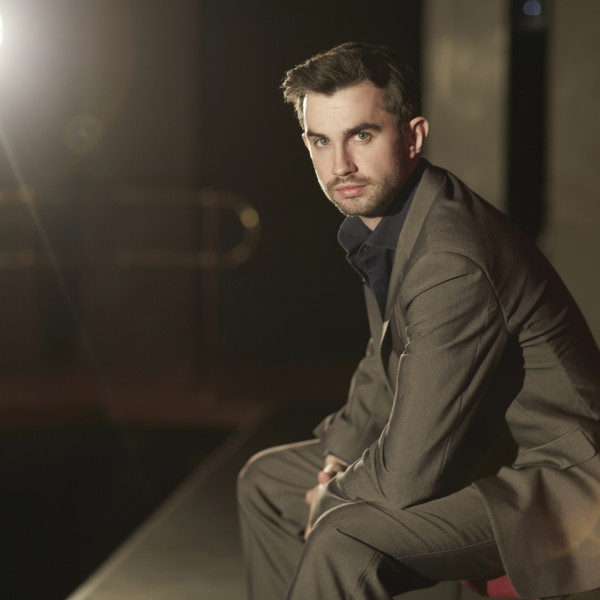 Tenor Anthony Gregory sits onstage, facing the camera against a backdrop of bright stage light.