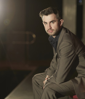 Tenor Anthony Gregory sits onstage, facing the camera against a backdrop of bright stage light.
