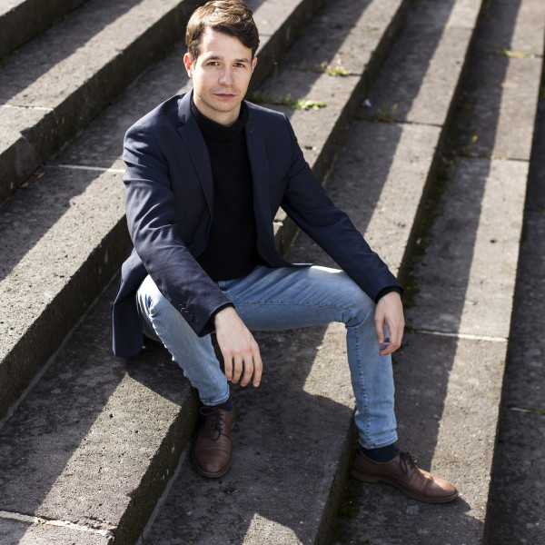 Bass-baritone Florian Störtz sits outdoors on large steps, arms resting on his knees as he looks up at the camera.