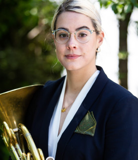 Horn player Lauren Reeve-Rawlings holds her horn in front of her, looking directly at the camera with a gentle smile.