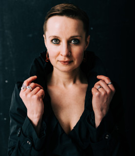 Mezzo-soprano Katie Bray looks directly into the camera, framing her face with both hands holding the collar.