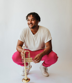 Aaron Azunda Akugbo squats down, holding his trumpet, laughing.