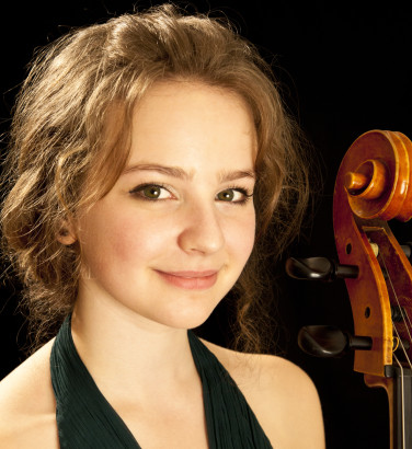 A woman in a dark dress looks towards the camera at a slight angle. She looks with a closed smile and is against a back background. The scroll of a cello is visible.